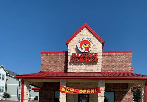 A brick building with a red roof and sign reading "Tasty's Chicken" with a banner that says "Now Open!" against a blue sky.