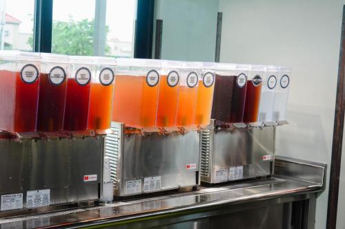 A row of beverage dispensers filled with various colorful drinks, set against a bright window.