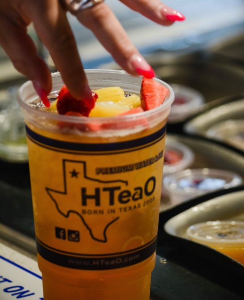 A hand adds fresh fruit to a large cup of iced tea from HTeaO, featuring a Texas-themed logo.