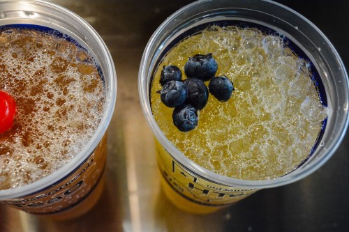 Two clear cups with fizzy drinks, one topped with a cherry and the other with blueberries.