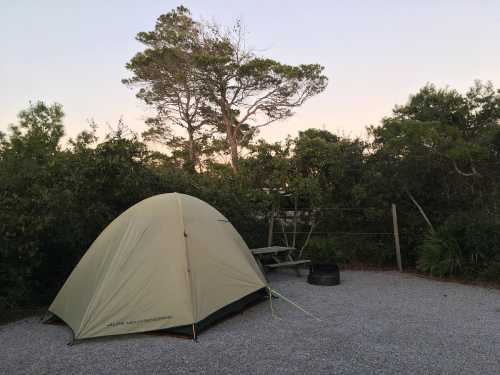 A green tent set up at a campsite surrounded by trees and gravel, with a picnic table and fire pit nearby.