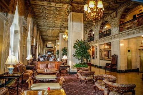 Elegant hotel lobby with ornate decor, plush seating, and large windows, featuring chandeliers and lush plants.