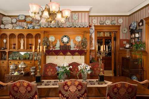 A vintage dining room with ornate decor, a wooden table, floral arrangements, and shelves filled with decorative plates.