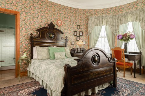 Cozy bedroom with floral wallpaper, a wooden bed, vintage decor, and a small desk by a window.