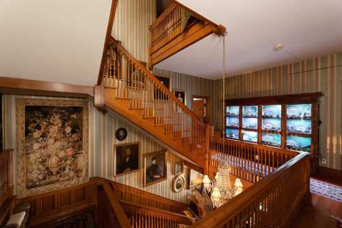 A wooden staircase with intricate railings, surrounded by vintage decor and framed portraits in a warmly lit hallway.