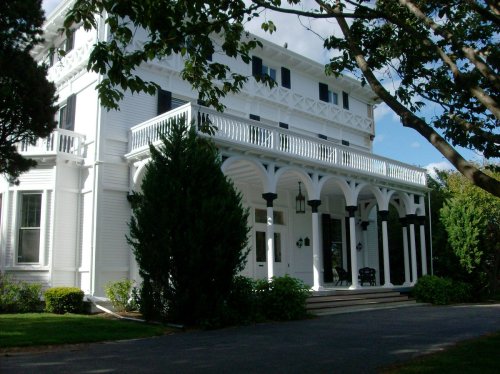 A large white house with a porch, black shutters, and lush greenery surrounding it on a sunny day.