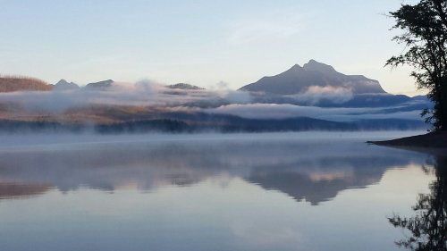 Serene lake scene with mist, mountains in the background, and a calm reflection on the water's surface.