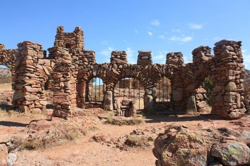 Ruins of a stone structure with arches and a gated entrance, set against a clear blue sky and rocky terrain.