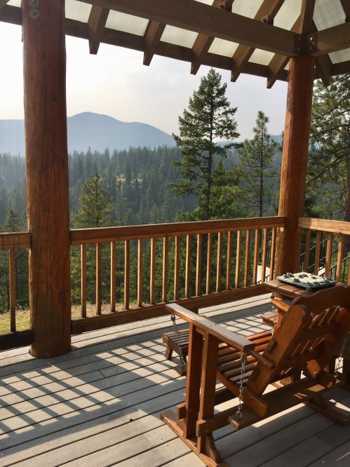 A wooden deck with a rocking chair overlooks a misty mountain landscape surrounded by tall trees.