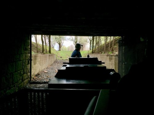 A person stands in a tunnel, framed by trees and light, with a tank-like structure in the foreground.