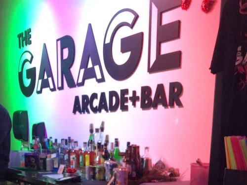 Colorful sign for "The Garage Arcade + Bar" with a bar area and various bottles in the foreground.