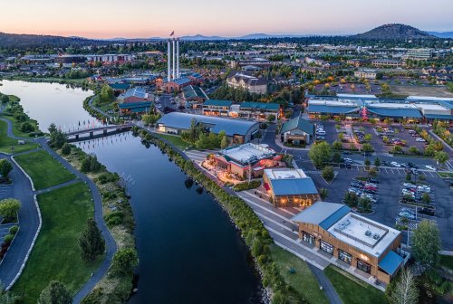 Aerial view of a vibrant riverside area with shops, restaurants, and a sunset backdrop in a scenic landscape.