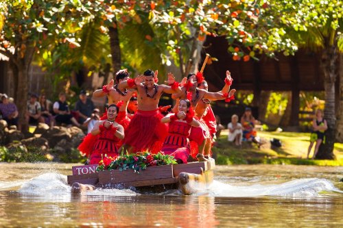 A group of dancers in traditional attire perform on a boat, surrounded by lush greenery and an audience by the shore.