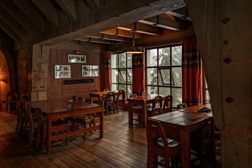 Cozy dining area with wooden tables, chairs, and large windows showcasing a view of trees outside. Warm, inviting atmosphere.