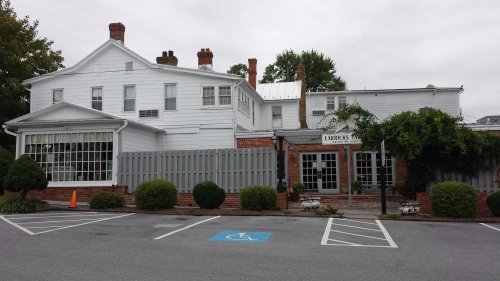 A white two-story building with a brick facade, surrounded by greenery and a parking lot.