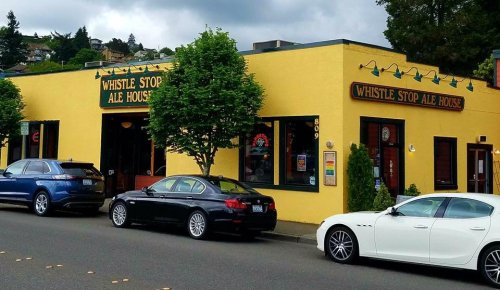 Exterior of Whistle Stop Ale House, a yellow building with signage, parked cars in front, and a cloudy sky above.