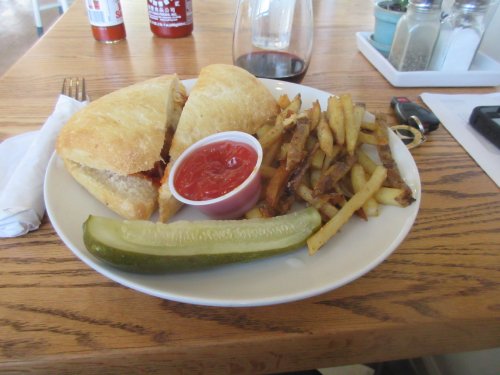 A plate with a sandwich, fries with ketchup, a pickle, and a glass of red wine on a wooden table.