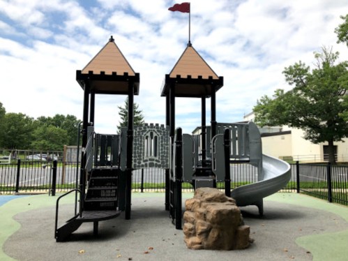 Playground with two towers, a slide, and a rock feature, set against a cloudy sky and green trees.