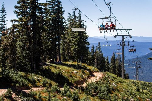 A chairlift carries four people over a forested mountain landscape on a clear day.