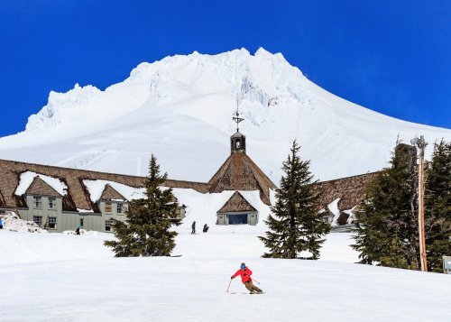 A skier glides down a snowy slope with a mountain peak and a rustic lodge in the background under a clear blue sky.