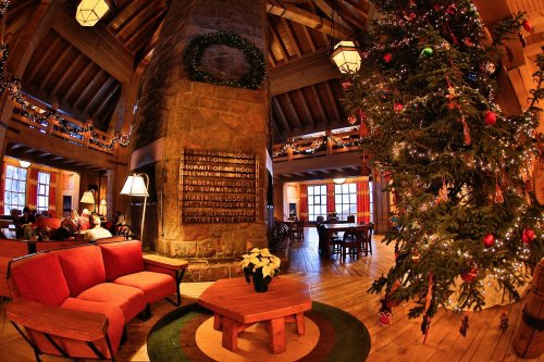 Cozy lodge interior with a large Christmas tree, festive decorations, and comfortable seating around a stone fireplace.