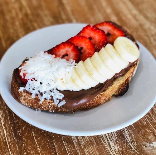 A slice of toast topped with chocolate spread, banana slices, strawberries, and shredded coconut on a white plate.