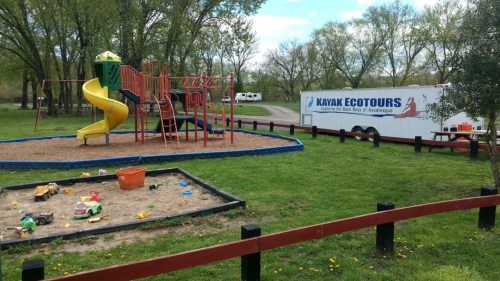 A playground with a slide and sandbox, next to a trailer for Kayak Ecotours, set in a grassy park area.