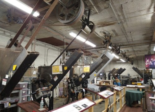A vintage printing press room with large machines and overhead pulleys, showcasing industrial equipment and displays.