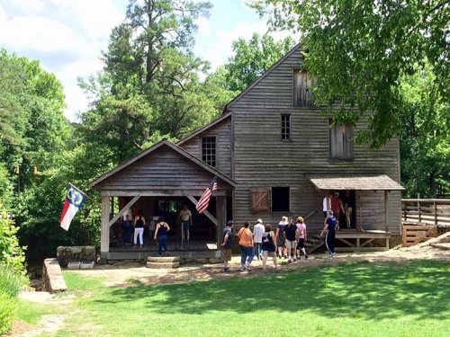 A historic wooden building surrounded by trees, with people gathered outside and flags displayed.