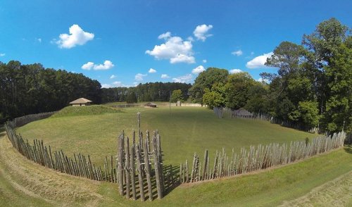 A grassy area enclosed by wooden palisades, surrounded by trees and under a blue sky with fluffy clouds.