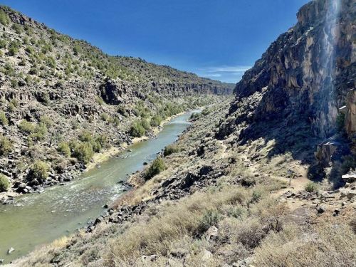 A winding river flows through a rocky canyon, surrounded by green vegetation and steep cliffs under a clear blue sky.