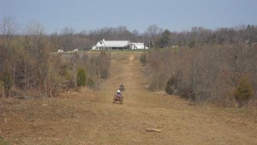 Two ATVs ride down a dirt path lined with trees, leading to a distant white building on a hill.
