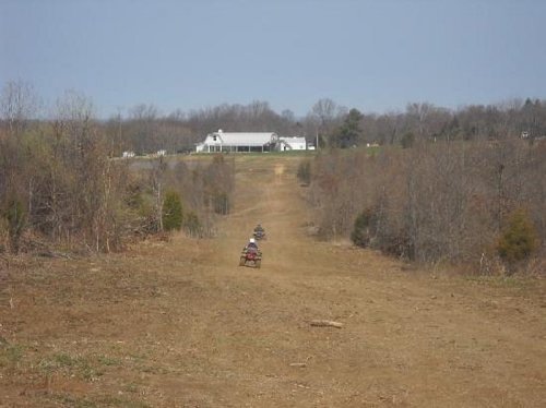 Two all-terrain vehicles ride down a dirt path through a wooded area, with a white house visible in the distance.