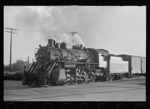 A vintage steam locomotive with smoke billowing, parked beside a freight car on a railway track.