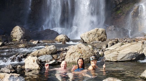 Three people relax in a natural hot spring, surrounded by rocks and a waterfall in the background.