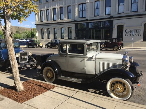 A vintage silver car parked on a city street near a historic building, with other classic cars in the background.