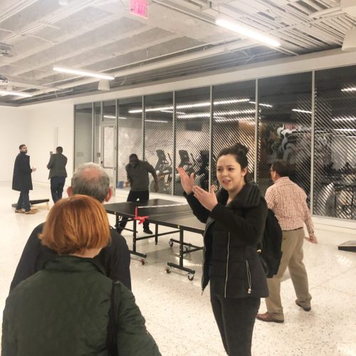 A group of people in an office space, with one woman gesturing near a ping pong table.
