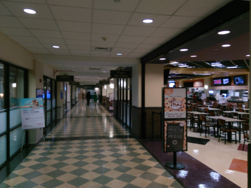A hallway in a food court with various dining options and empty tables, leading to a restaurant area.