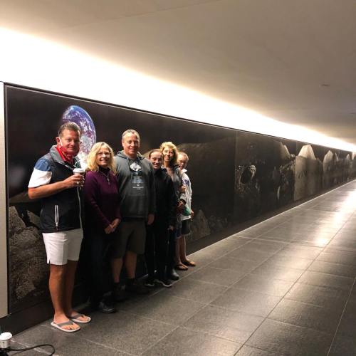 A group of five people stands in front of a large mural in a well-lit hallway, smiling and posing for the photo.