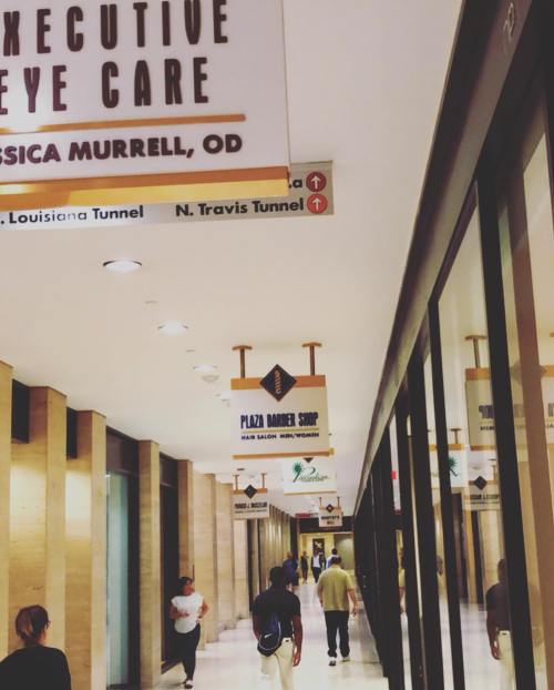 A hallway with signs for various businesses, including eye care and a barber shop, with people walking through.