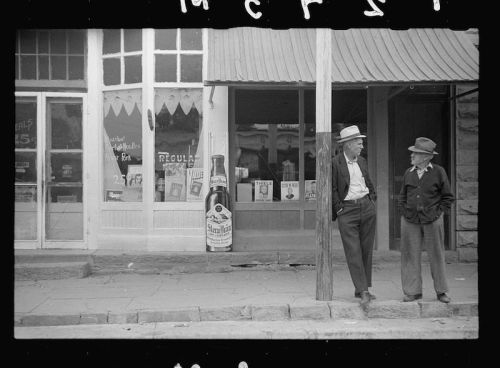 Two men in hats stand by a wooden post outside a storefront with vintage advertisements and a large bottle display.