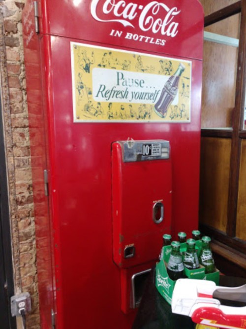 Vintage red Coca-Cola vending machine with a sign reading "Pause Refresh yourself" and bottles of soda in front.