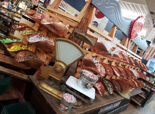 A display of colorful candies in wooden bins, with a vintage scale and metal buckets in a shop setting.