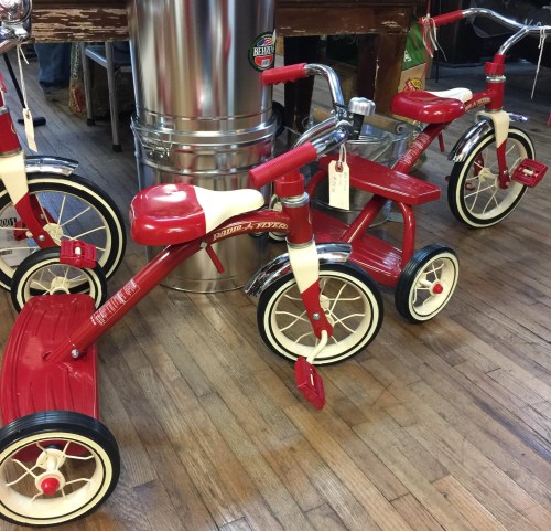 Three vintage red tricycles with white accents displayed on a wooden floor, featuring a metal can in the background.
