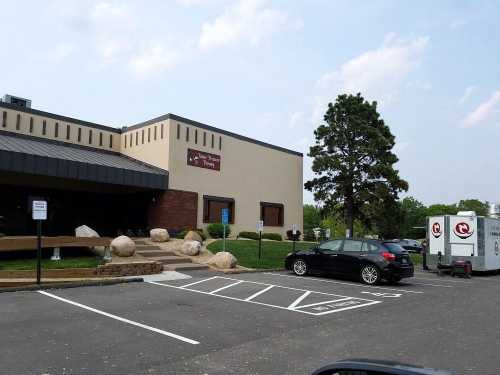 A modern building with a sign, surrounded by greenery and large rocks, featuring a parked car and a trailer in the lot.