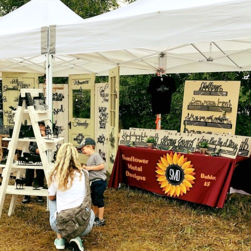 A vendor booth at an outdoor market featuring metal designs, with children exploring the displays and a woman taking photos.