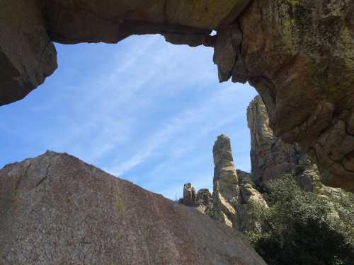 A rocky landscape framed by a natural stone arch, with blue sky and towering rock formations in the background.