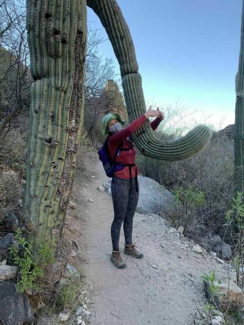 A person in hiking gear poses next to a large cactus on a trail, surrounded by desert vegetation.