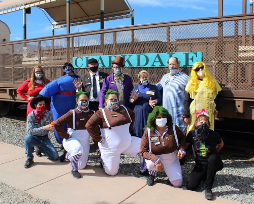 A group of people in colorful costumes and masks poses in front of a train, showcasing a festive atmosphere.