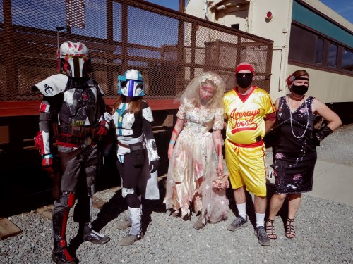 Five people in costumes, including a Mandalorian, a bride, and a retro sports outfit, posing near a train.
